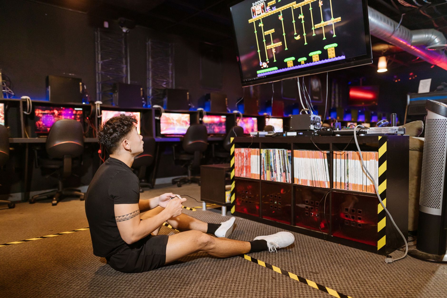 Homme assis par terre jouant intensément à un jeu vidéo dans une salle d'arcade moderne avec du matériel informatique vibrant.