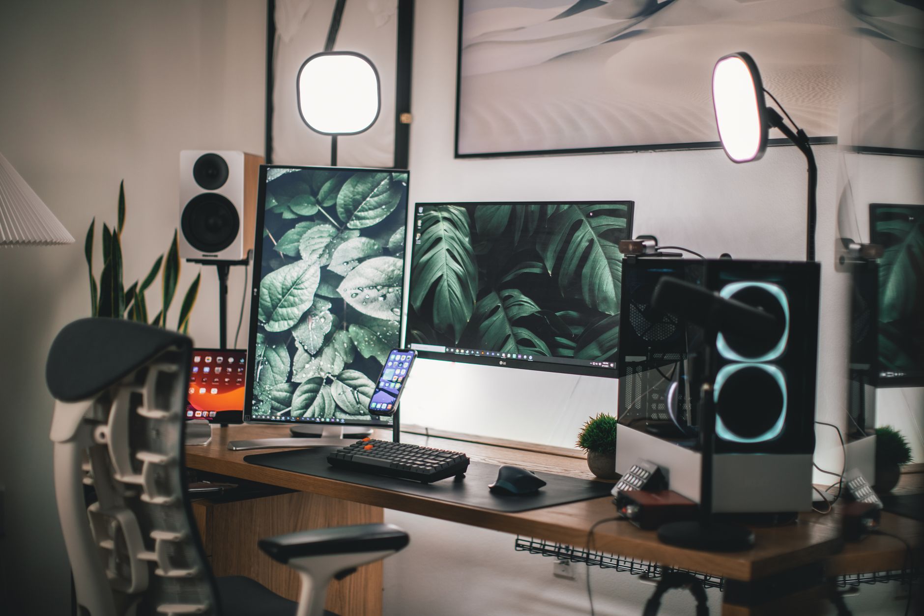 Bureau en bois avec deux écrans, clavier, souris, smartphone, enceinte, boîtier de PC lumineux, micro et lampes, devant une chaise ergonomique.