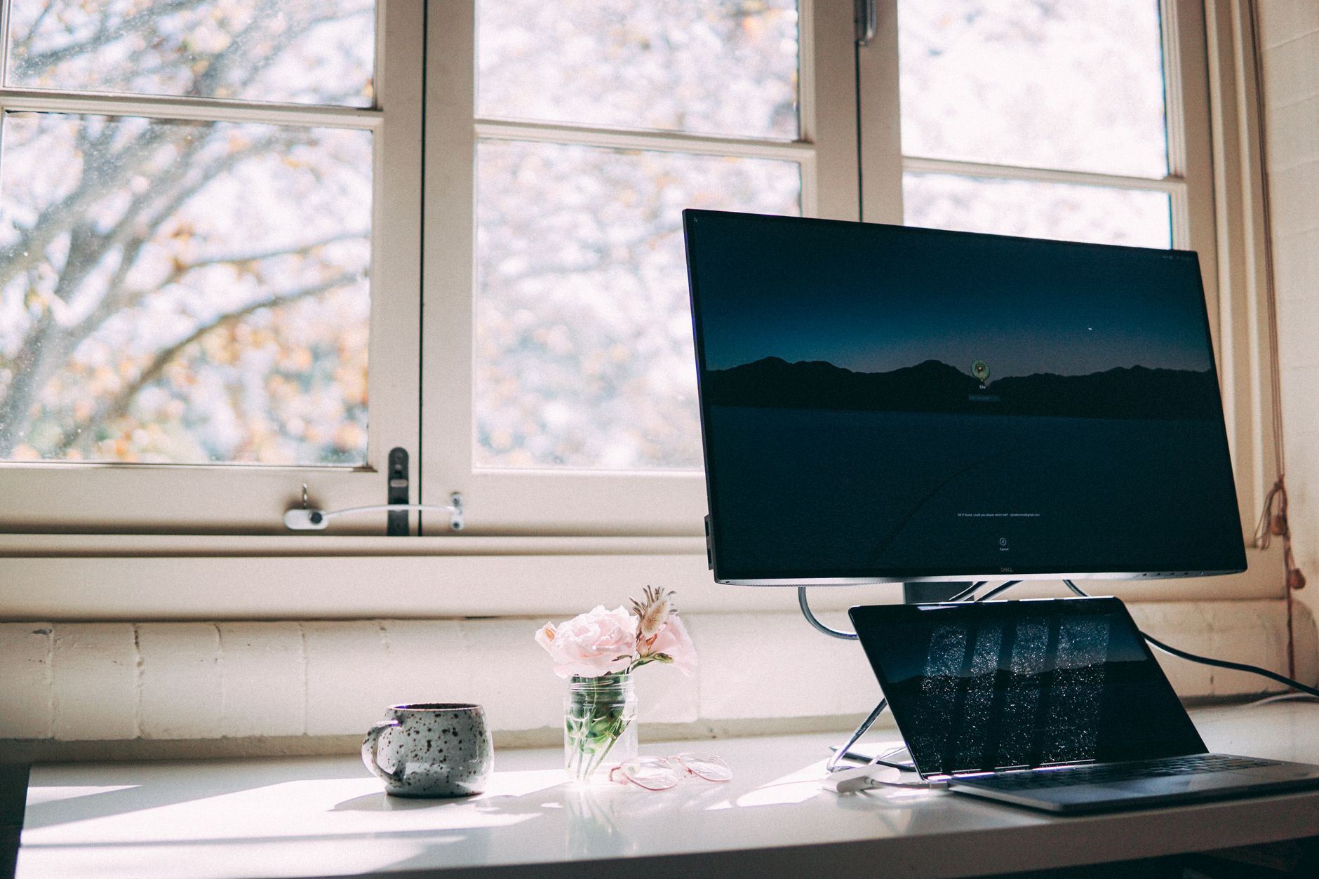 Bureau près d’une fenêtre avec un grand écran, un ordinateur portable ouvert, une tasse mouchetée et un petit vase de roses.