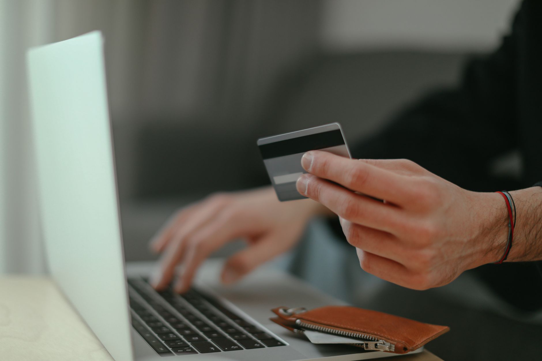 Une personne tient une carte bancaire d’une main et tape sur le clavier d’un ordinateur portable de l’autre, près d’un porte-monnaie en cuir.